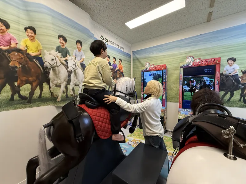 말 그림 벽화가 있는 체험실에서 선생님과 함께 승마 시뮬레이터를 타는 아이